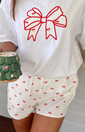 Girl wearing cherry print pajama shorts and white tee holding a hot chocolate mug.