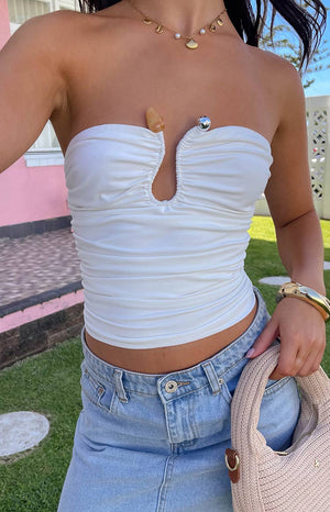 Model is wearing a white strapless top with a metal bar structuring the bust. Top has an orange stone detailing. Model is wearing gold jewellery and lightwash denim.