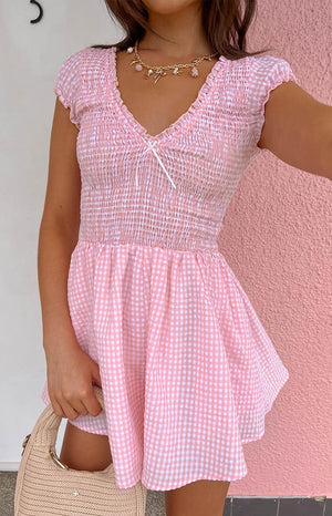 Model is wearing a pink gingham playsuit with ruffles. Model is wearing a gold and pink necklace and holding a woven handbag.